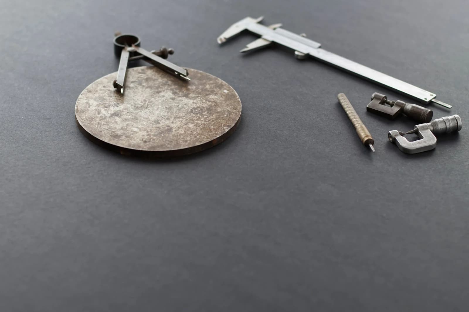 An undocumented machined steel disc next to a vernier caliper, micrometer and divider compass on a dark surface